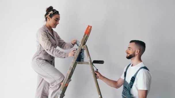 Жена стоит на лестнице, пока ее муж рисует стену. — стоковое видео