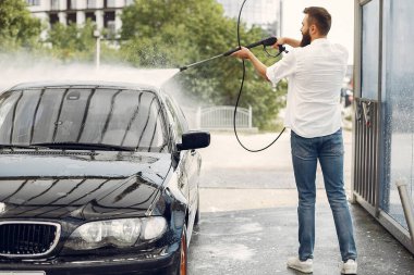Beyaz gömlekli bir adam arabasını yıkıyor.