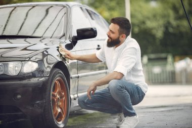 Beyaz gömlekli bir adam araba yıkamada araba siliyor.