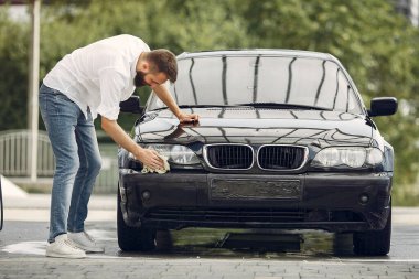 Beyaz gömlekli bir adam araba yıkamada araba siliyor.