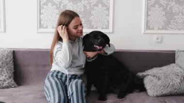 Esmer kız gündelik giysiler içinde beyaz odada siyah labradoru okşuyor kanepede oturuyor.