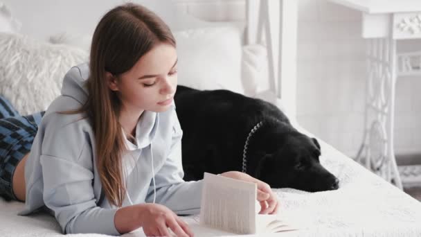 Brunette liggend op het bed en het lezen van een boek met een labrador — Stockvideo