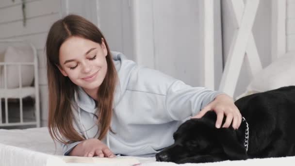 Brunett liggande på sängen och läser en bok med labrador — Stockvideo