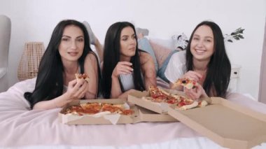 Üç muhteşem kız arkadaş yatakta uzanmış pizza yiyorlar.