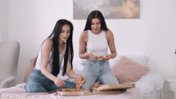 Dos chicas guapas de pelo oscuro comparten una pizza para llevar en la cama — Vídeo de stock