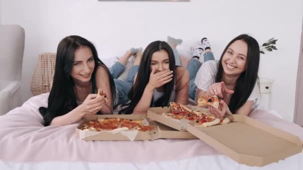 Три чудові подружки лежать на ліжку і їдять піцу — стокове відео