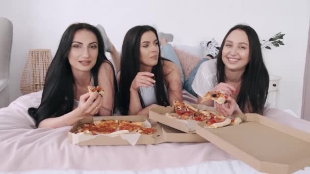 Drie prachtige vriendinnen liggen op bed en eten pizza — Stockvideo