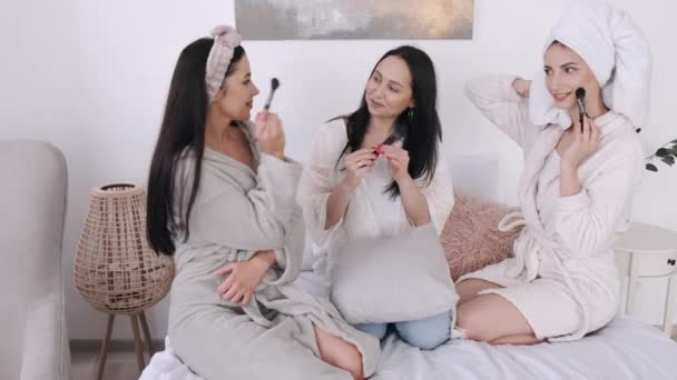 Jóvenes mujeres guapas están sentadas en la cama con pinceles de maquillaje y sonriendo — Vídeo de stock