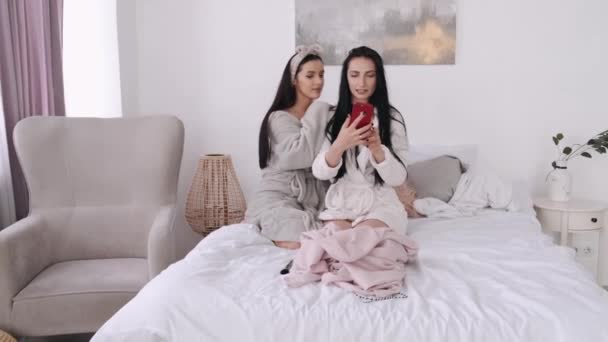 Two pretty girls are sitting on the bed and taking selfies — Stock Video