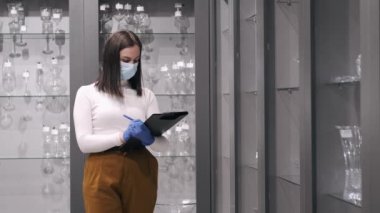 Maskeli ciddi kadın bir dükkandaki cam takımları kontrol ediyor.