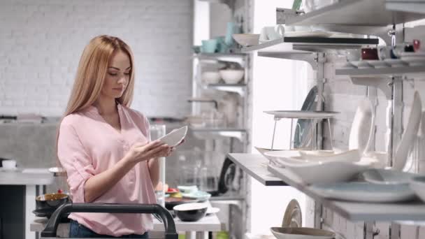 La bonita ama de casa está eligiendo vajilla de porcelana en la tienda de utensilios de cocina — Vídeo de stock