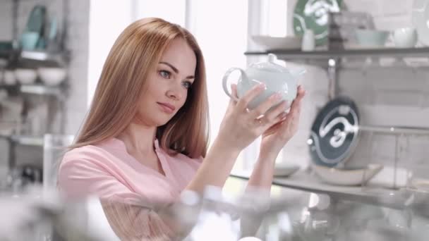 A jovem está olhando para o bule de vidro na loja de utensílios de mesa — Vídeo de Stock