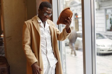 Pencerenin yanında duran kahverengi ceketli siyah adam.