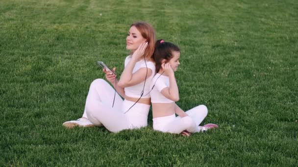 Mama i dziewczyna siedzą na trawniku i słuchają muzyki — Wideo stockowe