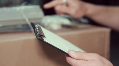 Fabrikadaki adam karton kutuları kontrol ediyor ve depodaki panoya bakıyor.