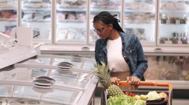 Elinde alışveriş arabası olan Afro-Amerikalı bir kadın donmuş ürünler reyonundan geçiyor.