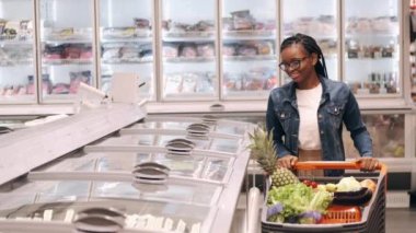 Elinde alışveriş arabası olan Afro-Amerikalı bir kadın donmuş ürünler reyonundan geçiyor.