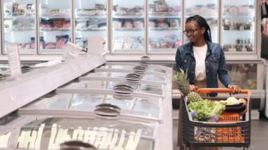 Elinde alışveriş arabası olan Afro-Amerikalı bir kadın donmuş ürünler reyonundan geçiyor.