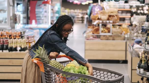 एक सुपरमार्केट में एक कार्ट में उत्पादों की जांच करने वाली अफ्रीकी लड़की — स्टॉक वीडियो