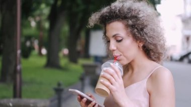 Çekici genç bayan kokteyl içiyor ve yaz şehrinde akıllı telefon kullanıyor.
