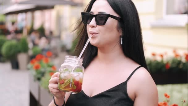 Wanita muda yang menarik berkacamata hitam meminum koktail di kota musim panas — Stok Video