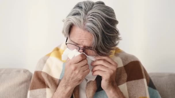 Un hombre mayor envuelto en manta sentado en un sofá y estornudando — Vídeos de Stock
