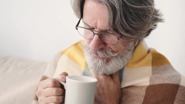 Senior man wrapped in blanket drinking mixture sitting at home — Stock Video