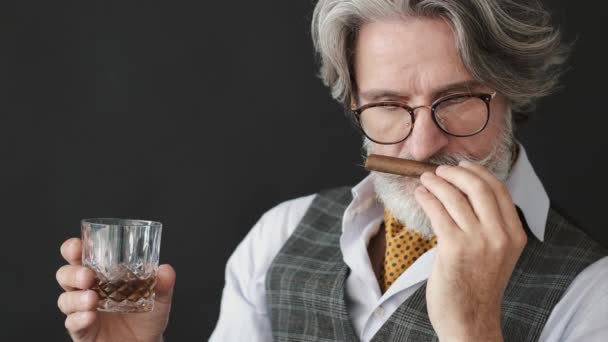 Homem maduro com um copo de uísque cheirando charuto sobre fundo preto — Vídeo de Stock