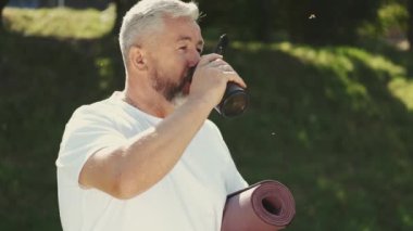 Kıdemli adam su içiyor ve dışarıda egzersiz paspası tutuyor.