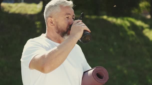 Senior homem água potável e segurando tapete de exercício fora — Vídeo de Stock