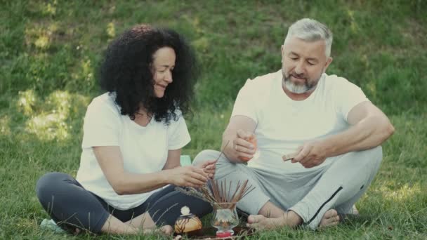 Healthy man and woman are sitting on the grass and preparing for meditation aroma sticks — Stock Video