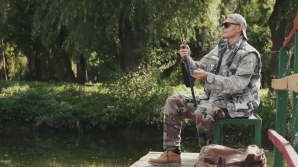 Mladý muž ve speciální uniformě a sluneční brýle rybaření na molu u jezera — Stock video