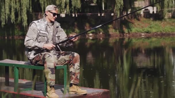 Ung man i en speciell uniform och solglasögon fiske på en brygga vid sjön — Stockvideo