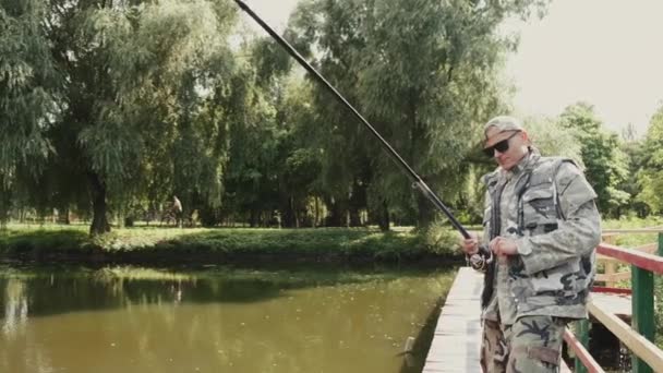 Man in special uniform fishing on a wooden pier in a park — Stock Video
