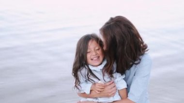 Anne ve gülümseyen kızı deniz kenarında sarılıyorlar.