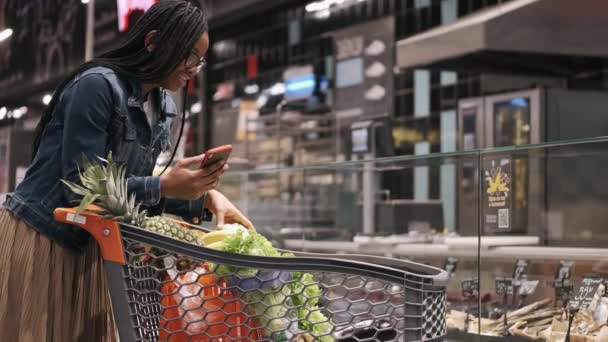Africana america chica con teléfono y carro en un supermercado — Vídeo de stock