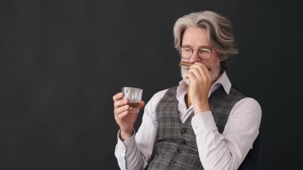Homem maduro com um copo de uísque cheirando charuto sobre fundo preto — Vídeo de Stock