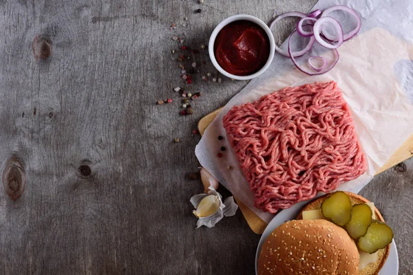 raw minced meat on paper, ingredients for burger with tomato, onion and seasonings on wooden background