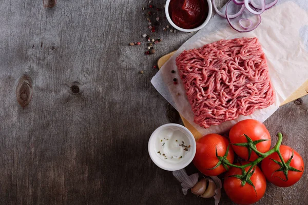 raw minced meat on paper, ingredients for burger with tomato, onion and seasonings on wooden background