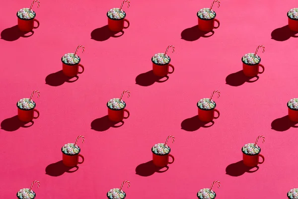 Red enamel mug full of marshmallows and candy cane on pink background. Hard light. Pattern