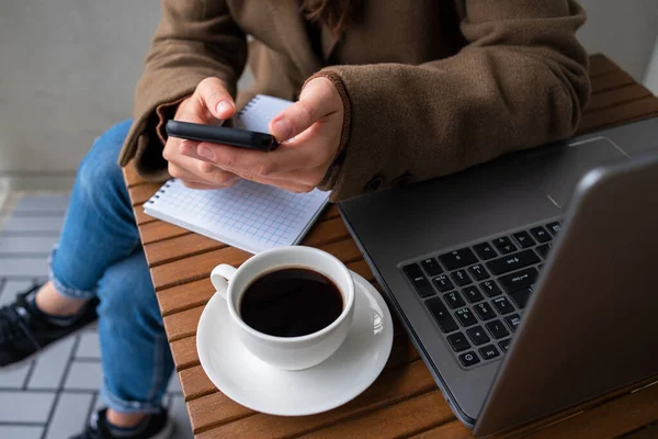 Woman in wool coat working in street cafe with laptop. Cup of coffee on wooden table. Using phone