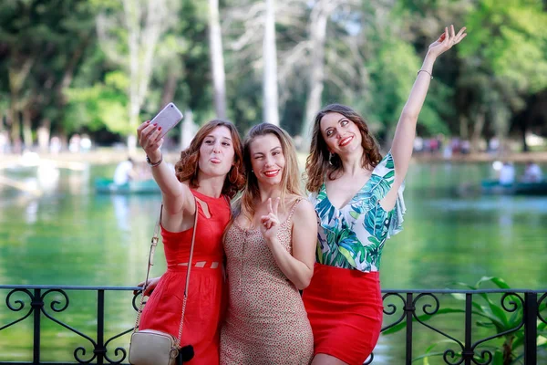Three girls, friends, smile and take a selfie together. The blonde girl winks and with her fingers makes the sign of victory. In the background the green lake in the public park.