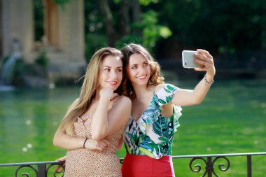 Two girls take a selfie together and smile. In the background the green lake in the outdoor park. clipart