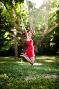 Mutlu kız sevinç için atlama ve kollarını yükseltmek. Arka plan ağaç ve yeşil ormanın Park. Çiller ve Kızıl saçlı, kırmızı kısa bir elbise giymiş kızla.