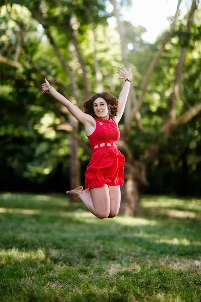 快乐的女孩跳跃的喜悦和举起她的胳膊 在背景树和绿色森林的公园 女孩与雀斑和红色头发 穿着短的红色礼服 — 图库照片