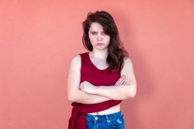 Sıradan giyinen esmer kız, kolsuz kırmızı tişört ve kot şort. Renkli bir arkaplanda yarım boy portre, yüz ifadesi somurtuyor.