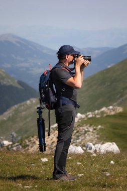 Bir dağın tepesinde duran yürüyüşçü bir kamera tutuyor ve çevresini çevreleyen manzaranın fotoğrafını çekiyor. Fotoğrafçı sırt çantası ve tripodla yürüyüş kıyafeti giyer..