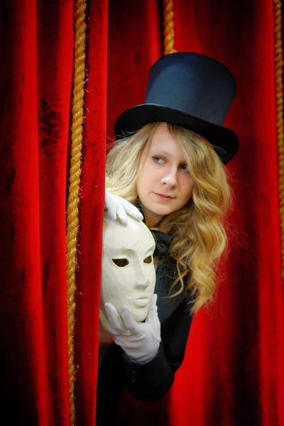 the actress with a theatrical mask stands near the red theater curtain