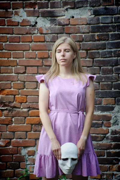 A blonde girl in a lilac dress walks through the ruins of an old brick building with a white theatrical mask in her hands