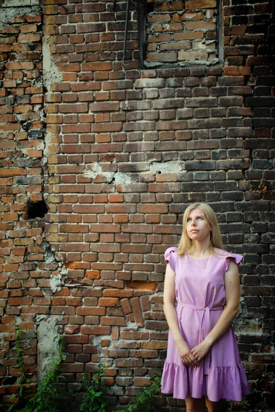 A blonde girl in a lilac dress walks through the ruins of an old brick building